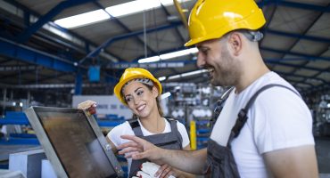 Factory worker explaining trainee how to operate industrial machine using new software on touch screen computer.