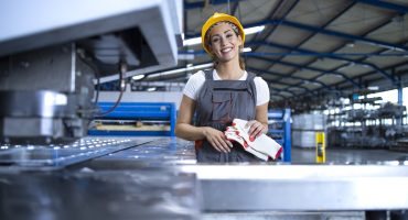 Portrait of female factory worker in protective uniform and hardhat standing by industrial machine at production line.